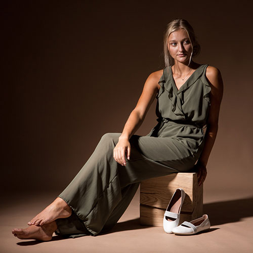 Junge Frau sitzt in einem olivgrünem Jumpsuit auf einem Hocker. Vor ihr stehen weiße Sommerschuhe von Marc.